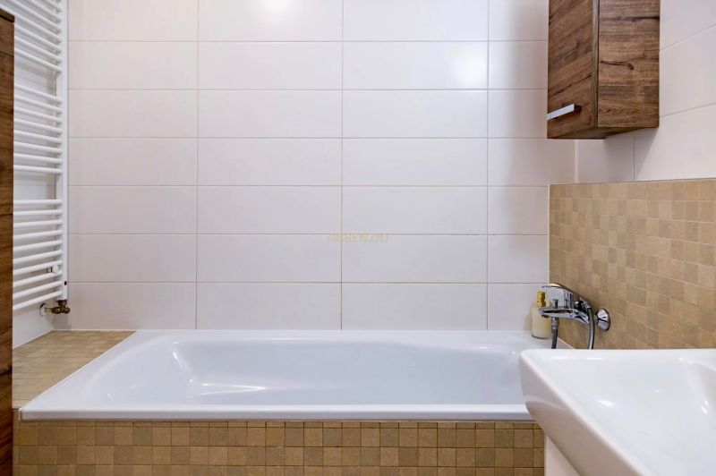 Bathroom in a 2-room apartment with white tiles and a bathtub, wooden decor on the cabinet.
