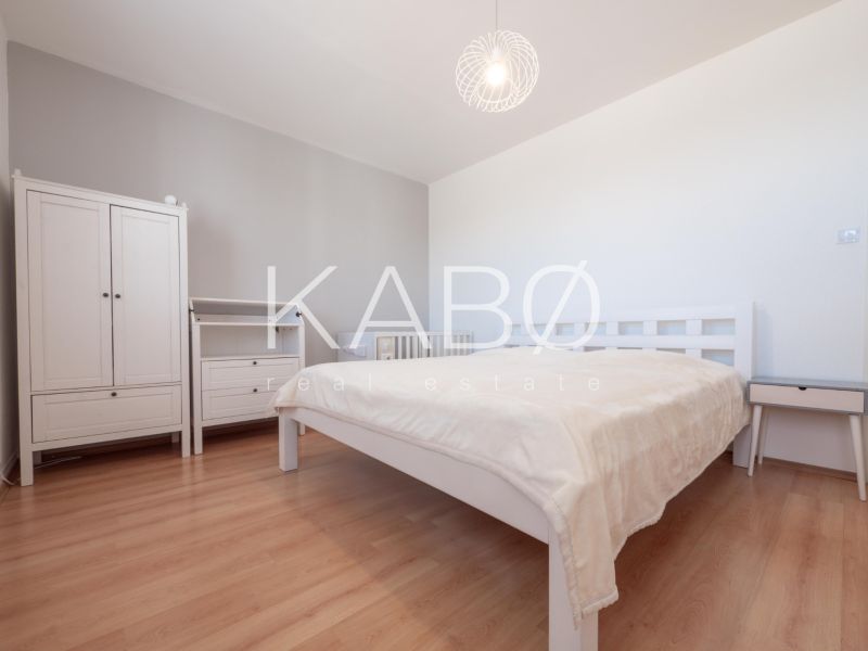Bedroom in a two-room apartment with wood-patterned flooring, light furniture, and a lamp.