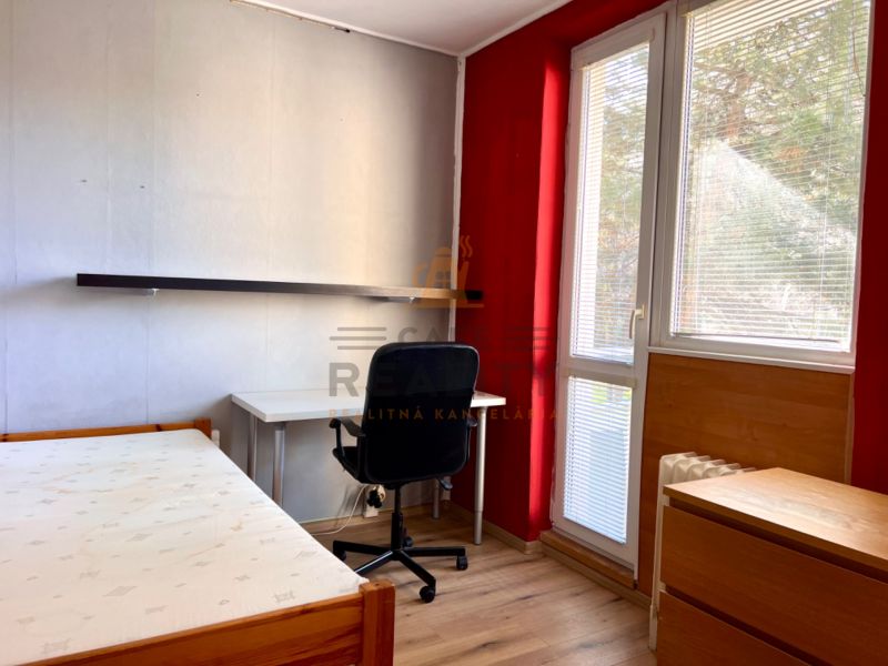 A room in a 2-room apartment with a wooden decor floor, a desk, and a bed.
