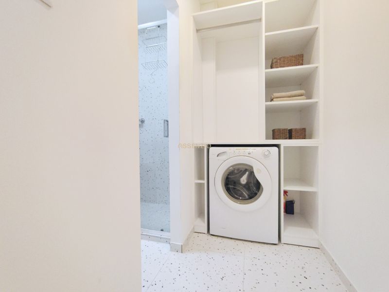 Bathroom with a shower and a washing machine in a 2-room apartment. White shelves increase storage options.