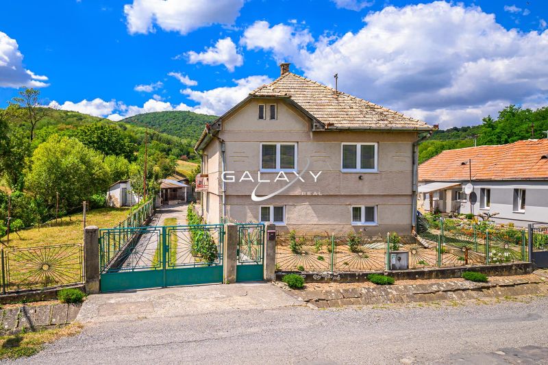 A family house in Hajnáčka surrounded by greenery and a fenced yard, with a blue sky.