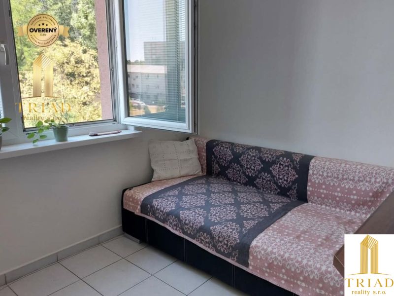 A sofa in the corner of the room with an open window and flowers on the windowsill.