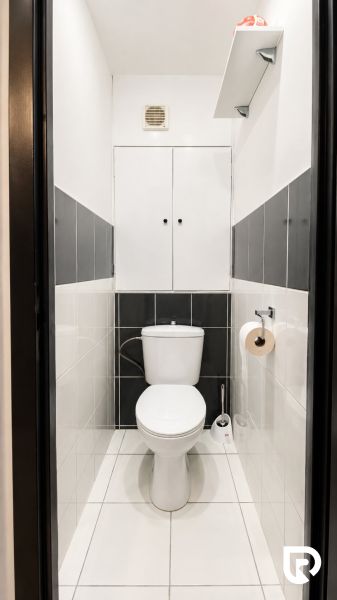 A bathroom with white and gray tiles, storage space above the toilet.