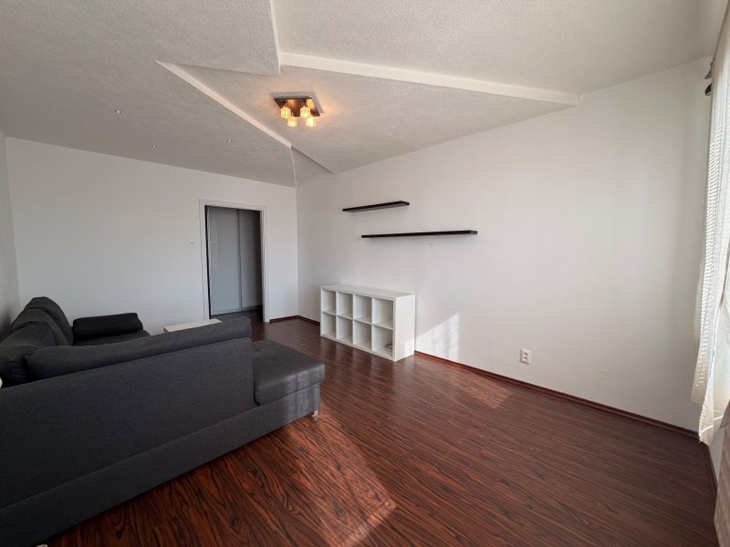Living room in a three-room apartment with a wooden decor floor, gray sofa, and shelves.