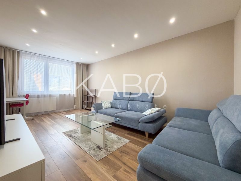 Living room in a 2-room apartment with a blue sofa, glass table, and wood-patterned flooring.