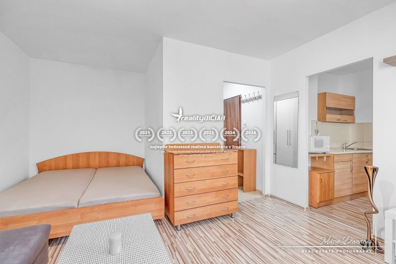 A studio apartment with a bed, a dresser, and a kitchenette with a wooden decor floor.