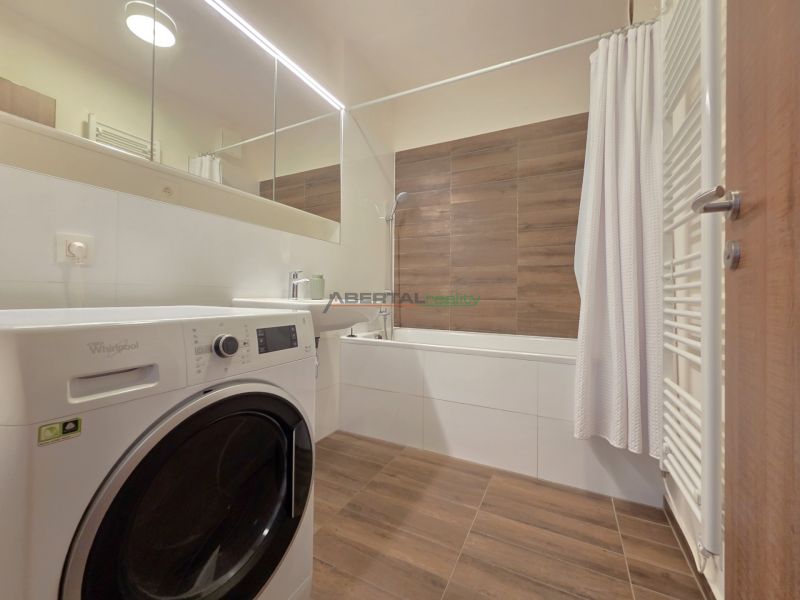 A bathroom in a 2-room apartment with a shower, washing machine, and a wooden decor floor.