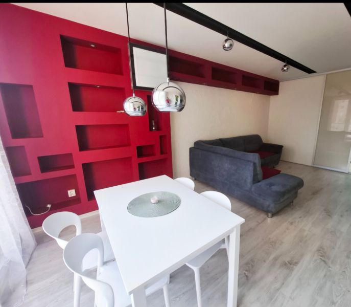 The interior of a 3-room apartment with red shelves, a white table, and a gray sofa.