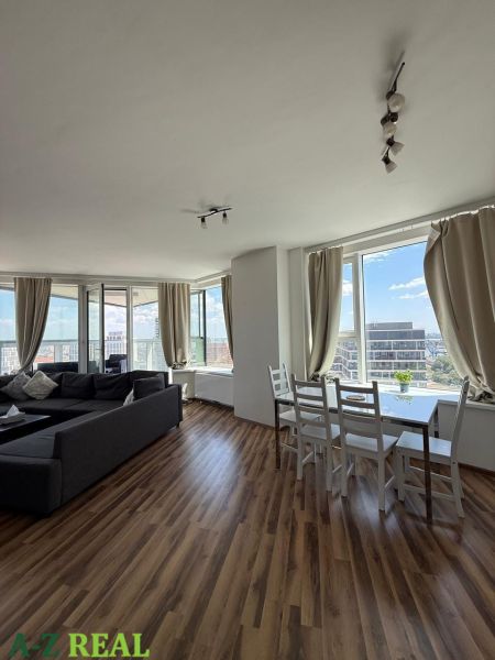 Living room in a three-room apartment with a wood-patterned floor and large windows.