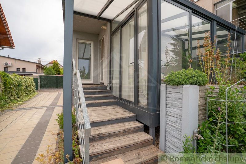 A cottage in Senec with a glazed veranda, staircase, and paved courtyard, bordered by greenery.