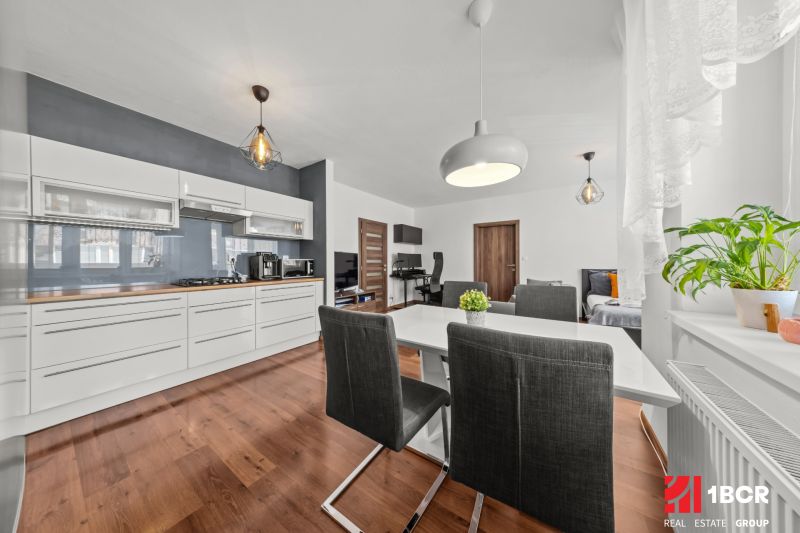 Living room of a two-room apartment with a kitchen, dining table, and wooden decor flooring.