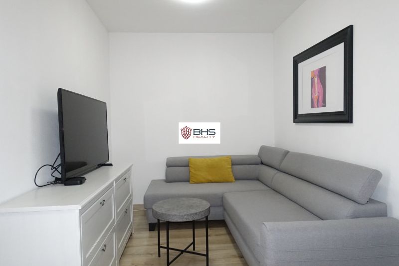 Living room in a two-room apartment with a sofa, television, and wooden decor flooring.