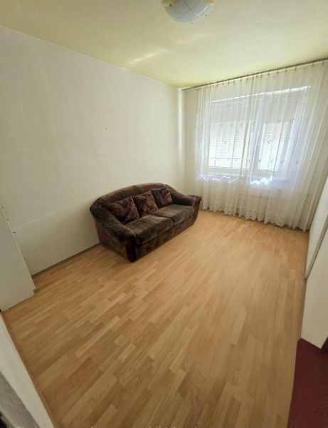 A room in a 3-room apartment with a sofa, wood-decorated flooring, and curtains.
