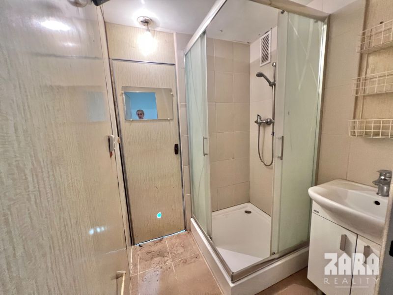 A bathroom with a corner shower in a 3-room apartment, a sink, and light tiles.