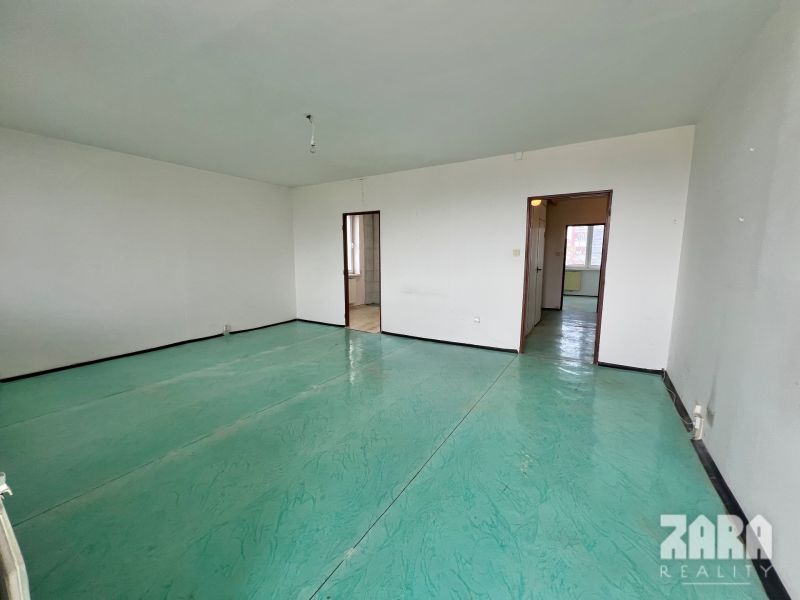 A spacious room of a 3-room apartment with white walls and a green floor.