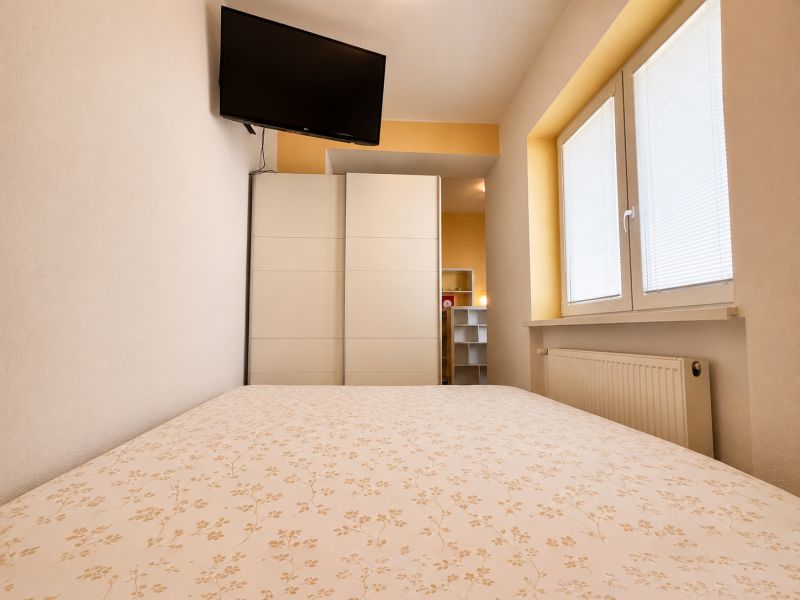 A bedroom in a studio apartment with a TV, a wardrobe, and a window with blinds.