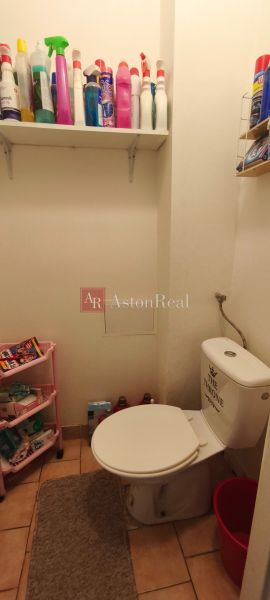 A toilet with beige tiles and a shelf with cleaning products in a studio apartment.