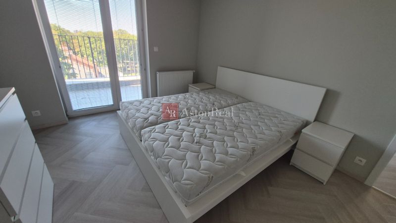 A bedroom in a three-room apartment with a wood-patterned floor and a large window.