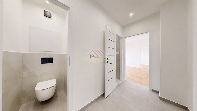 A toilet with ceramic tiles and a transition to a room with a wooden decor floor.
