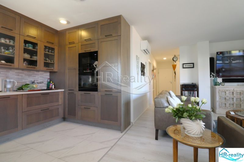 A kitchen in a holiday apartment with an open living room, wooden cabinets, and light flooring.