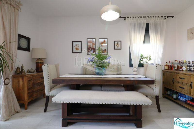 Dining room with a wooden table, upholstered chairs, decorative paintings, light curtains, wooden sideboard.