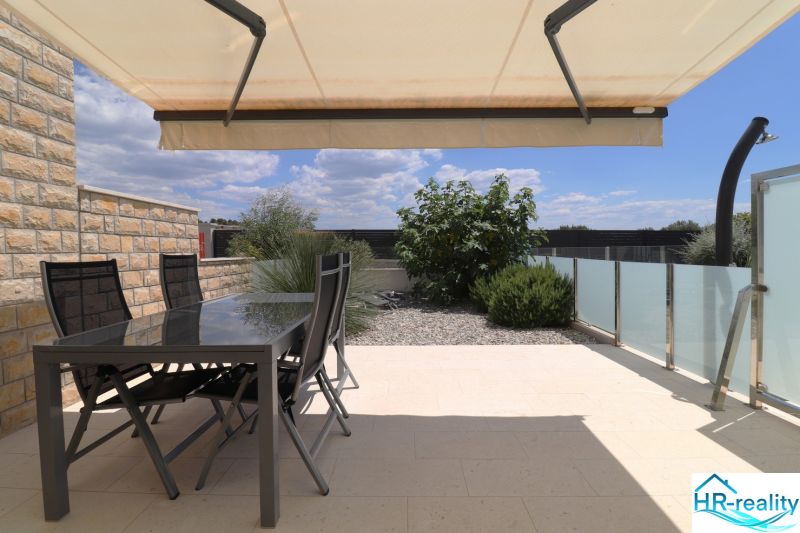 The terrace of the recreational apartment in Tribunj with spacious seating and an awning, surrounded by greenery.