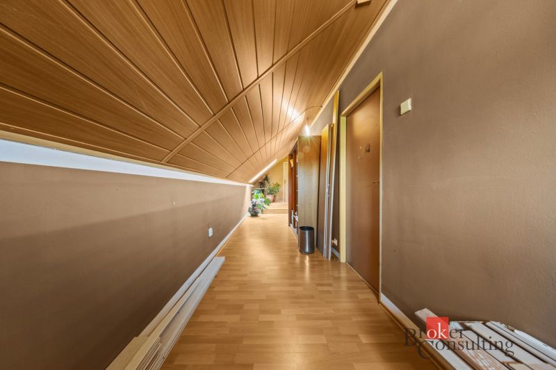 A hallway with a slanted ceiling, a floor with a wooden decor, a plant at the end.