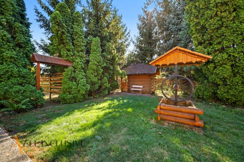 A garden with a wooden well, a gazebo, and a bench, surrounded by greenery near plots in Pezinok.