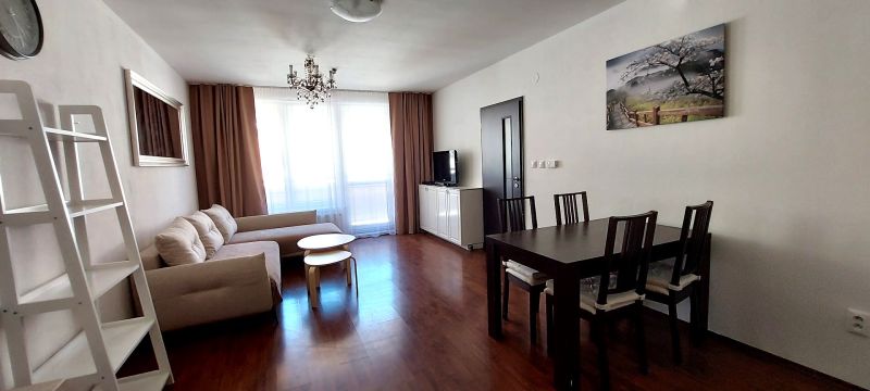 Living room of a three-room apartment with a sofa, dining table, and wood-patterned flooring.