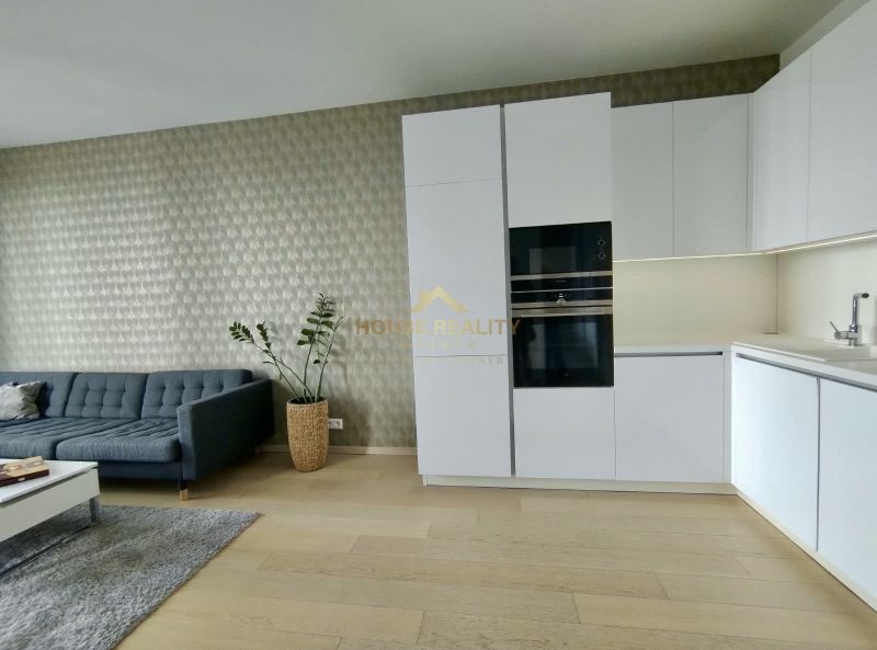 Living room in a two-room apartment with a sofa, modern kitchen, and wood-patterned flooring.