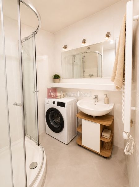 A bathroom in a 2-room apartment with a shower, washing machine, and sink.
