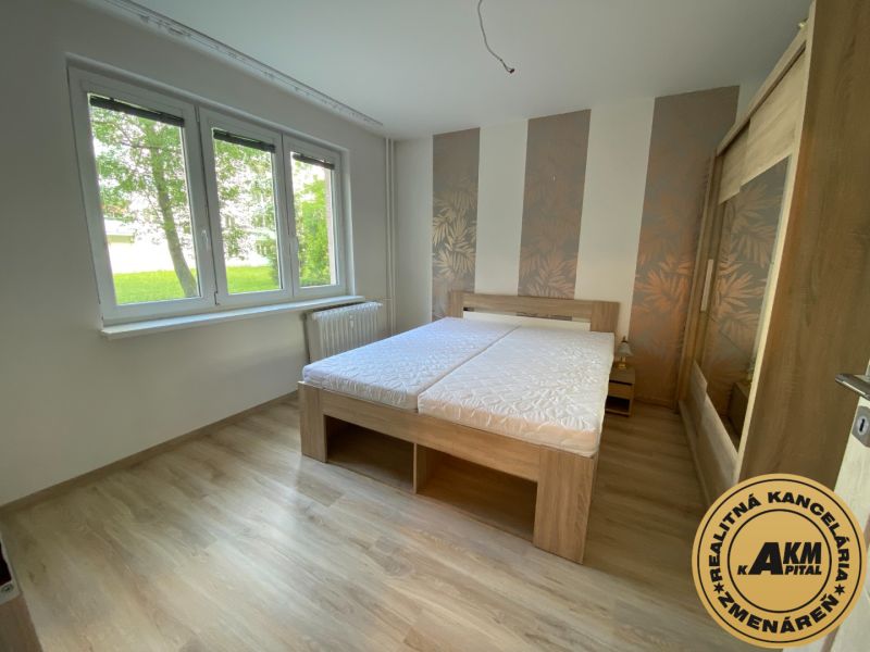 A bedroom in a two-room apartment with a wood-patterned floor and large windows.