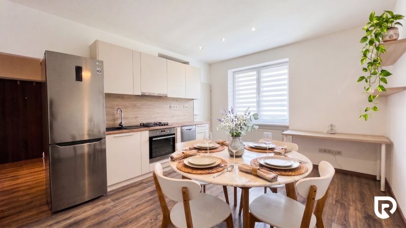 A kitchen of a two-room apartment with a wooden decor floor, a dining table, and appliances.