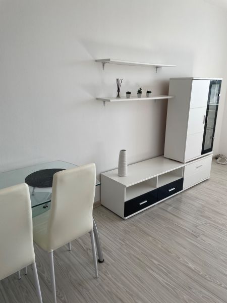 Studio apartment interior with wood-patterned flooring, white furniture, and a glass table.