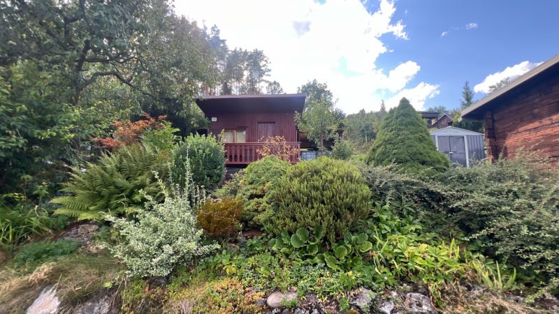 A wooden cabin in the green gardens of Ružomberok surrounded by trees and shrubs under a clear sky.