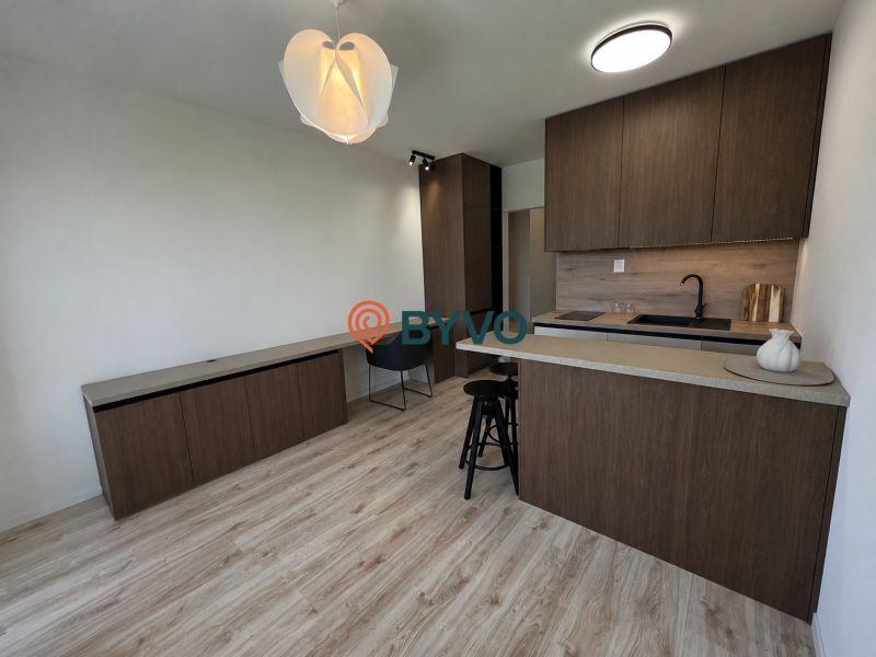 In the studio apartment, there is a kitchen unit with dark furniture and a floor with a wood design.