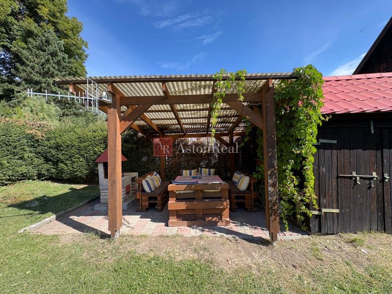 A wooden pergola in front of the cottage in Beňadiková surrounded by greenery and a sunny yard.