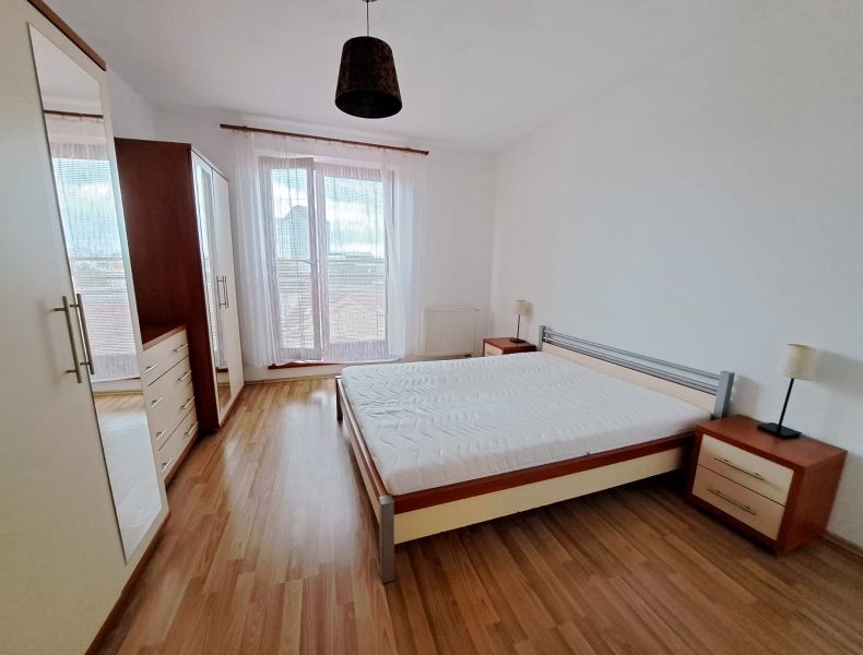 A bright bedroom in a two-room apartment with a wooden decor floor and large windows.