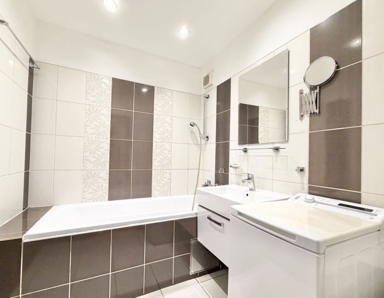 A bathroom in a 2-room apartment with a shower, sink, and washing machine, tiled.