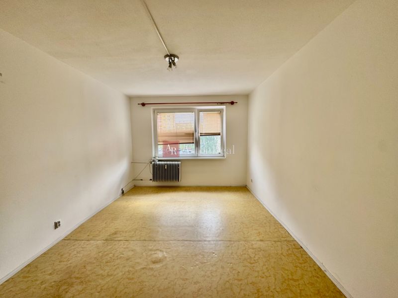 A room in a 2-room apartment with a beige floor and a window with a radiator.