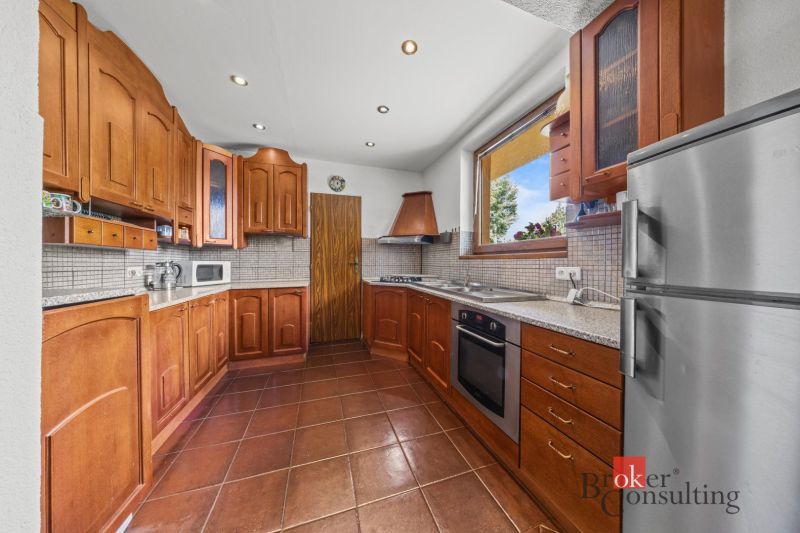 A kitchen in a family house with a ceramic floor, wooden cabinets, and a large window.