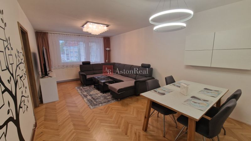 Living room with a corner sofa, dining table, and wood-patterned flooring in a three-room apartment.