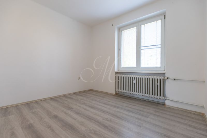 An empty room with a window, a wooden decor floor, and a radiator in a 3-room apartment.