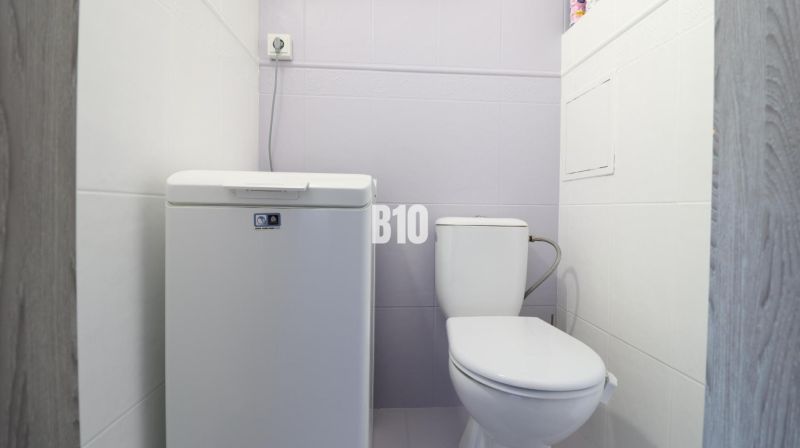A bathroom in a 3-room apartment with a toilet and a top-loading washing machine.