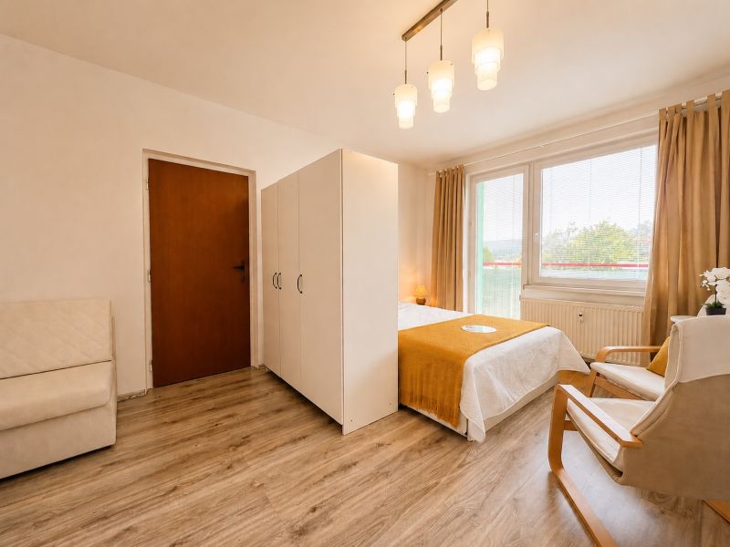 A one-room apartment with a wooden decor floor, a bed, and a chair by the window.