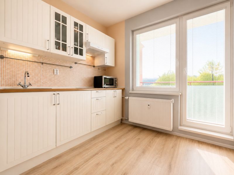 A kitchen in a one-room apartment with a wooden decor floor, large windows, and white cabinets.