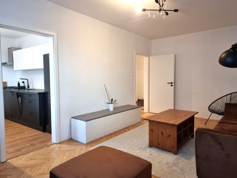 Living room in a two-room apartment with a wooden decor floor, visible into the kitchen.