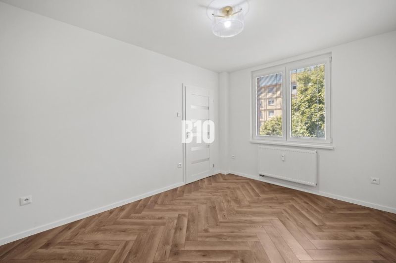 A room in a three-room apartment with a wood-patterned floor and a large window.