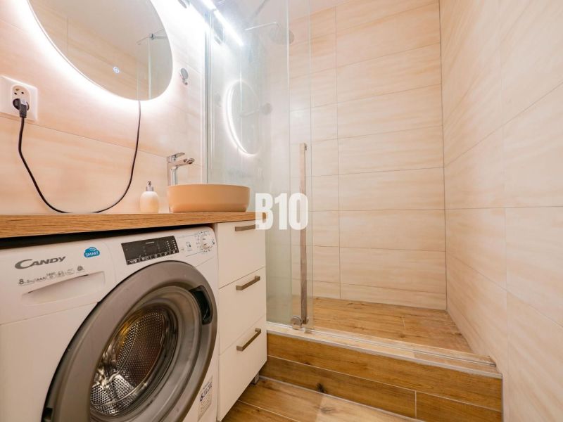 Bathroom in a studio apartment with a washing machine, mirror, and wooden decor flooring.