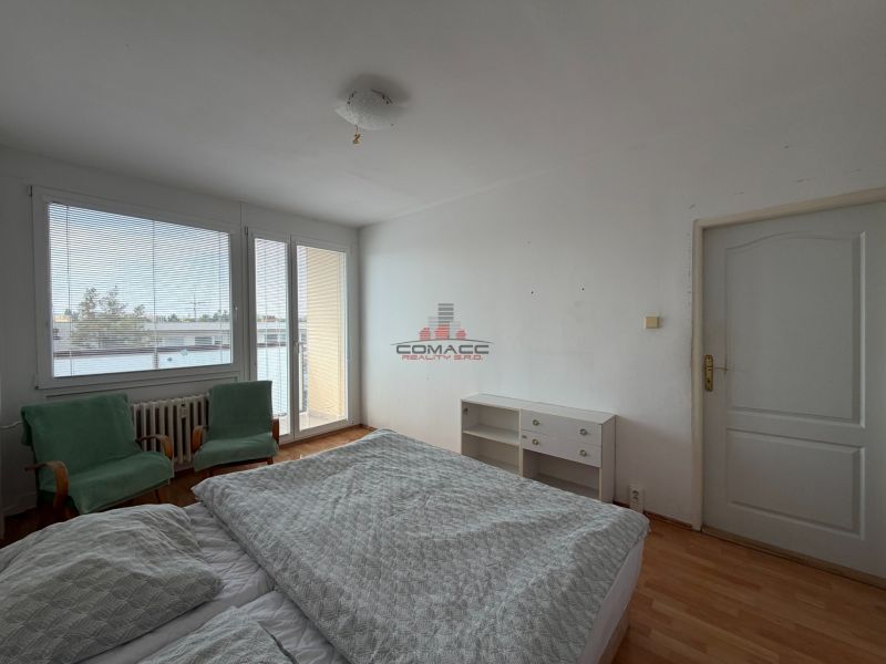 A bedroom in a 3-room apartment with a wood-patterned floor, a bed, and chairs.